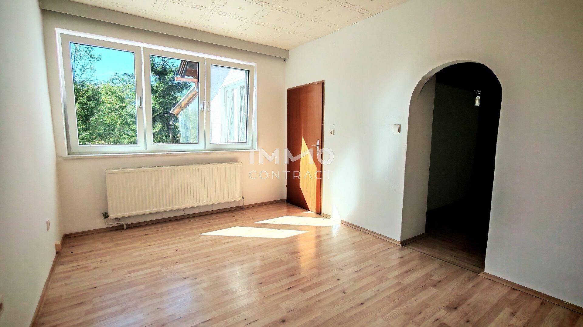 Helles Zimmer mit Parkettboden, großem Fenster und einem Rundbogen zum Flur.
