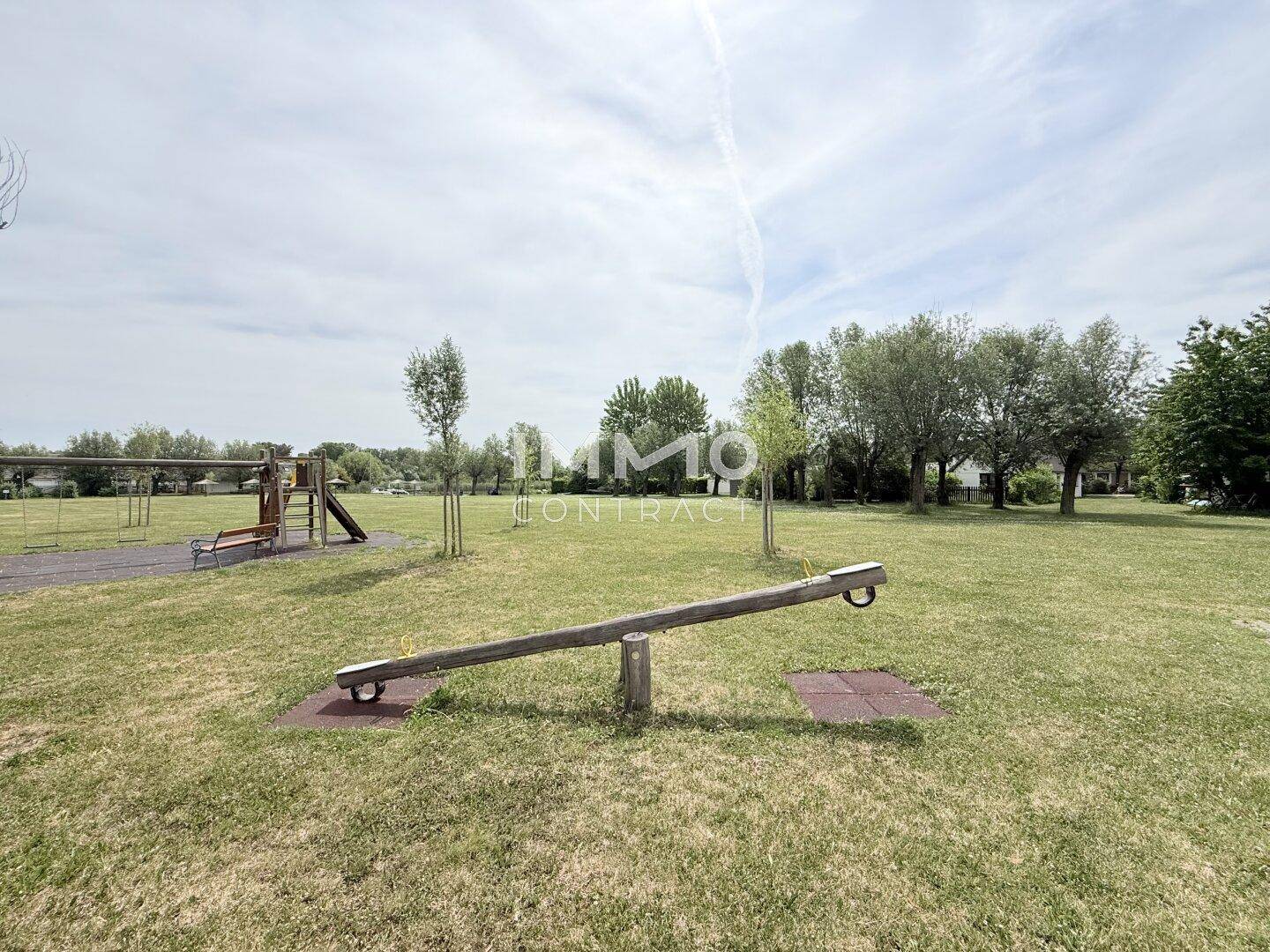 Kinderspielplatz mit einer Holzwippe und weiteren Spielgeräten auf einer großen Rasenfläche.