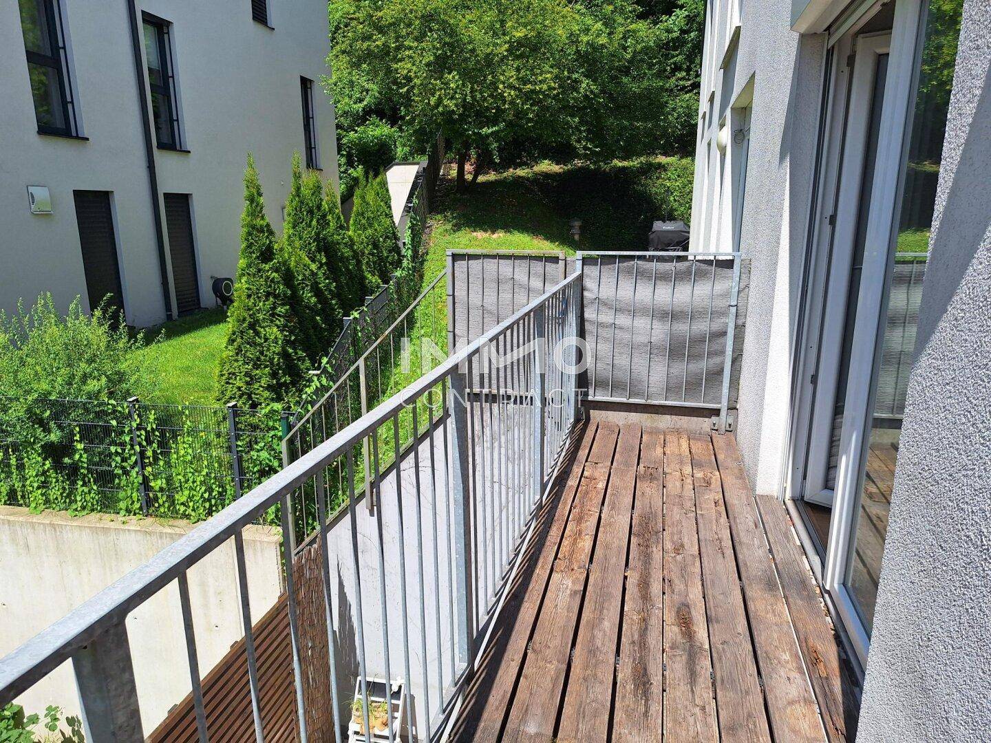 Kleiner Balkon mit Holzboden und Metallgeländer, umgeben von Bäumen und Grünflächen.