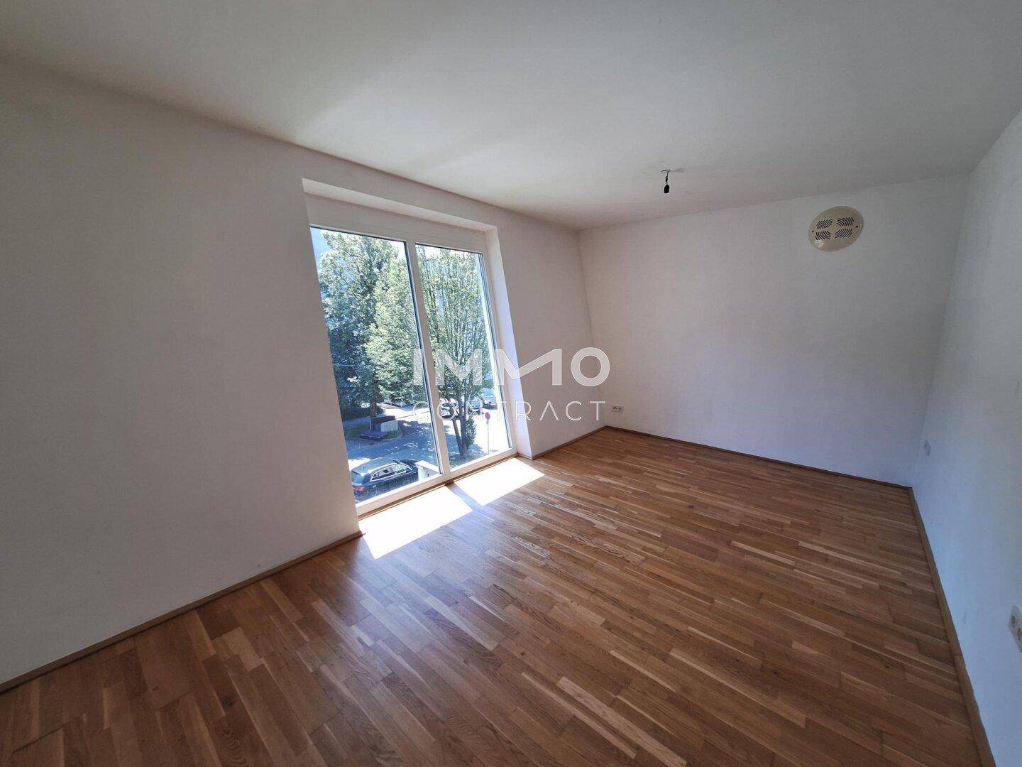 Helles Schlafzimmer mit Holzboden und großem Fenster, das viel Tageslicht hereinlässt.
