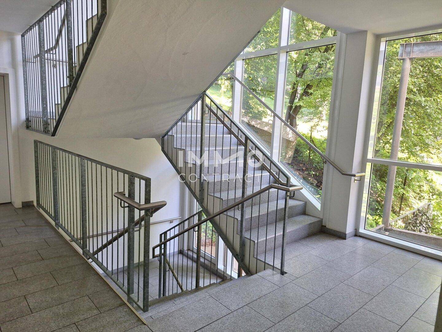 Helles Treppenhaus mit großen Fenstern, Metallgeländer und Blick ins Grüne.