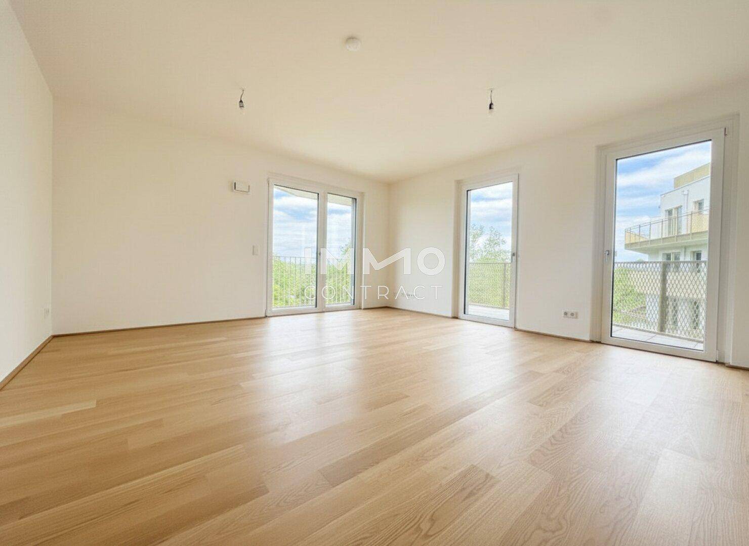 Helles, leeres Zimmer mit Holzboden und großen Fenstern, die viel natürliches Licht hereinlassen.