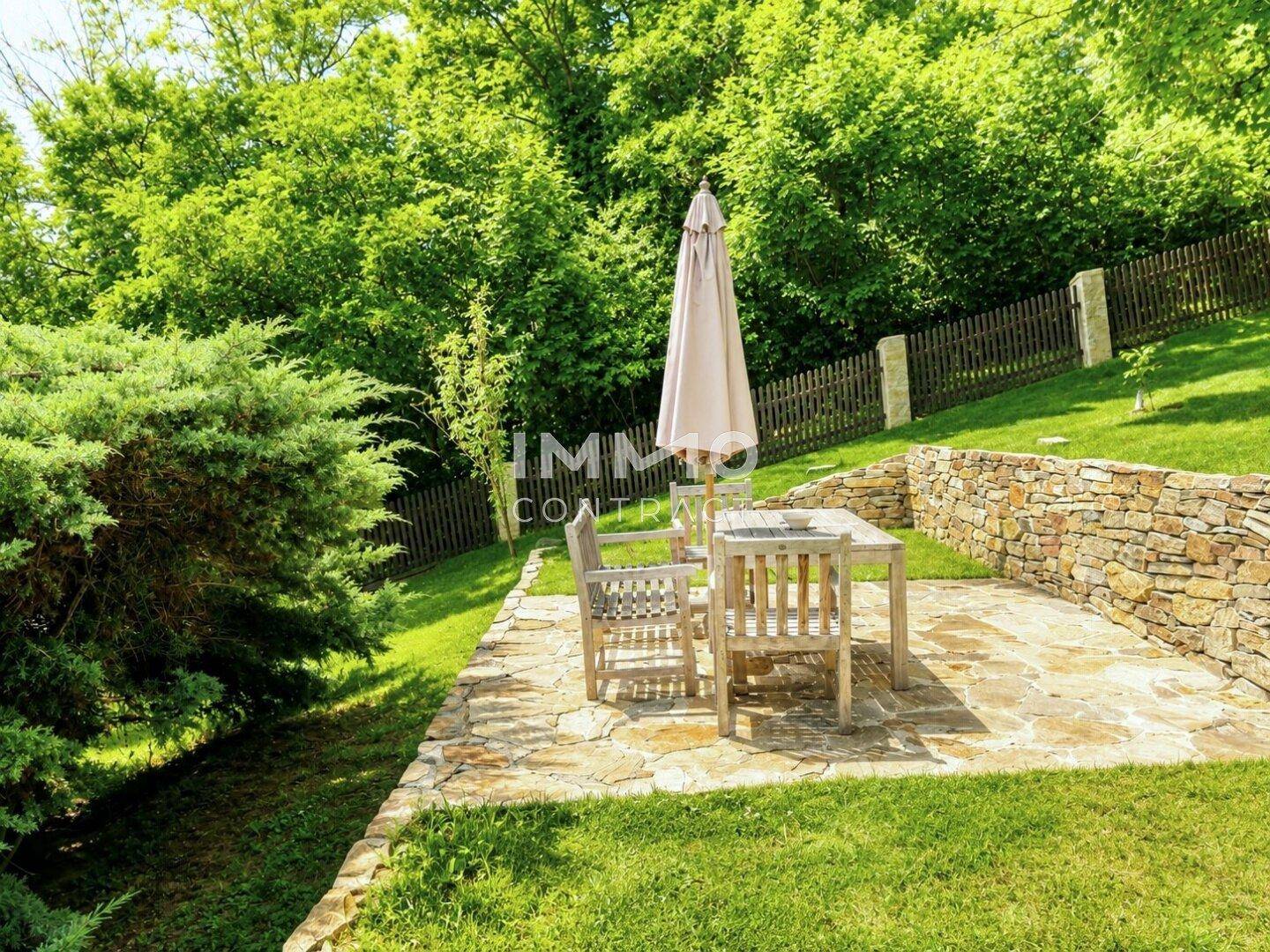 Gemütliche Terrasse im Garten mit Holztisch, Stühlen und Sonnenschirm, umgeben von Grün.