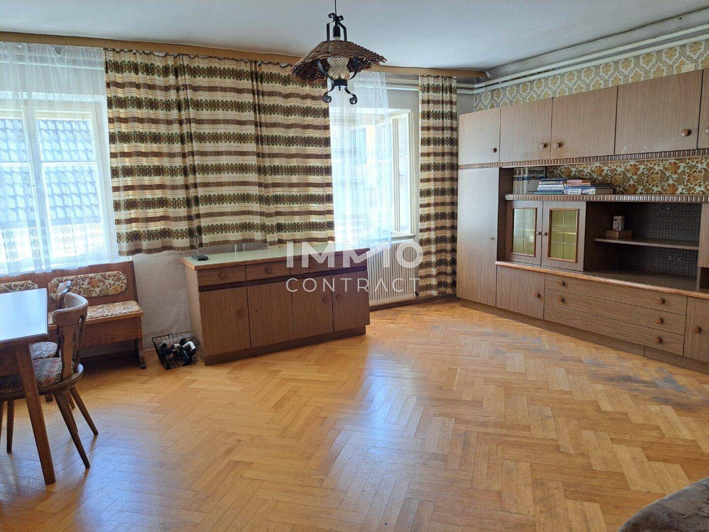 Wohnbereich mit Fischgrätparkett, Holzmöbeln und großen Fenstern mit Vorhängen.