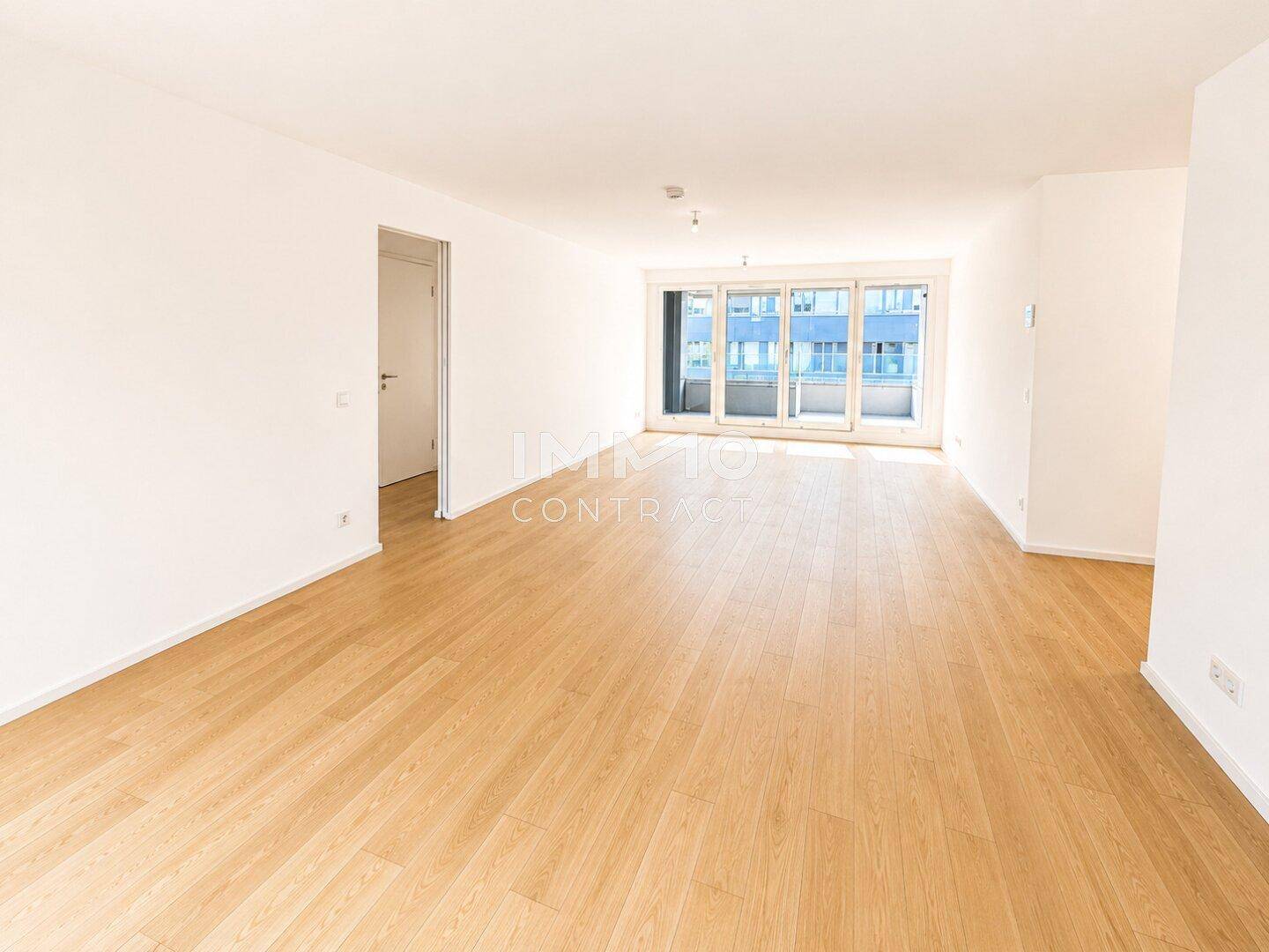 Heller, leerer Wohnraum mit Holzboden und großen Fenstern mit Blick auf die Umgebung.