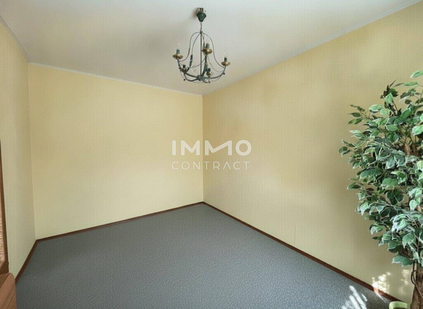 Leeres Zimmer mit hellgelben Wänden, grauem Teppichboden und einem Kronleuchter.