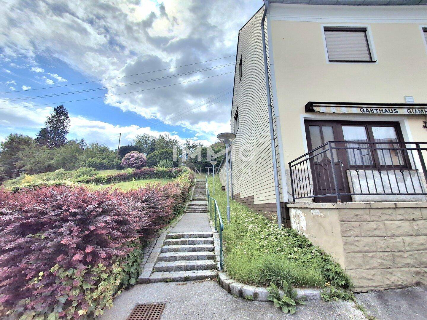 Seitenansicht des Gebäudes mit Außentreppe und üppiger Vegetation, die zum Garten führt.