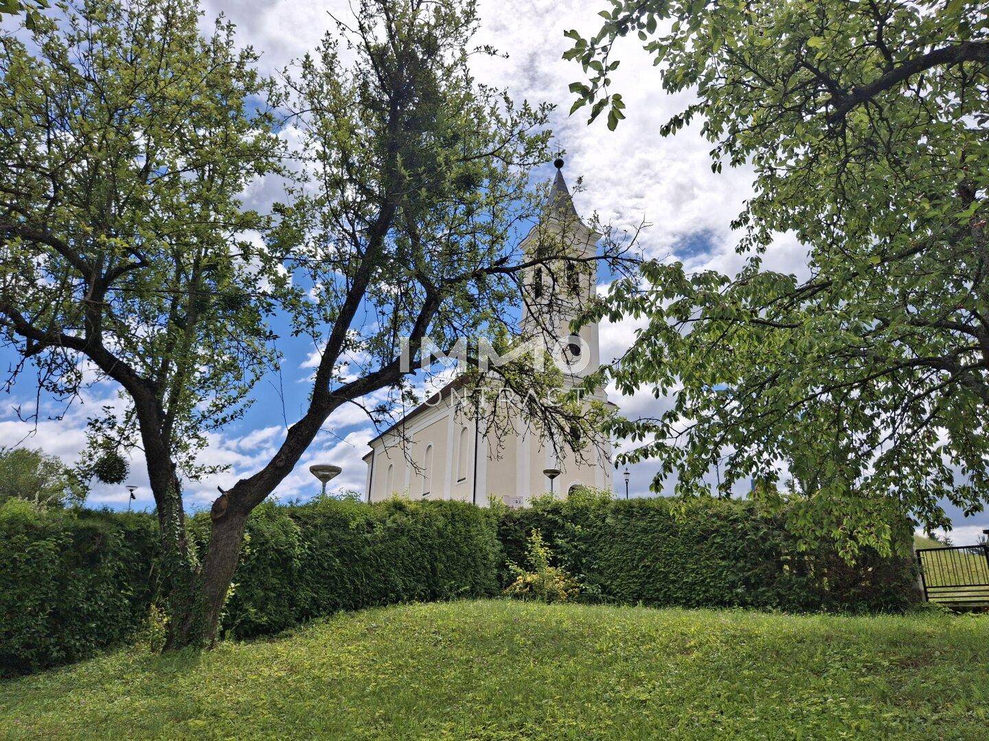 Historische Kirche mit hohem Kirchturm, umgeben von Bäumen und einer grünen Hecke.
