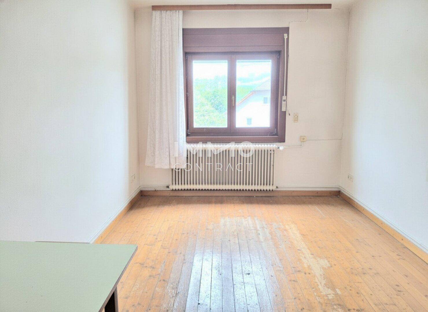 Schlafzimmer mit Holzdielenboden, einem Fenster und einem Heizkörper unter dem Fenster, einfach eingerichtet.