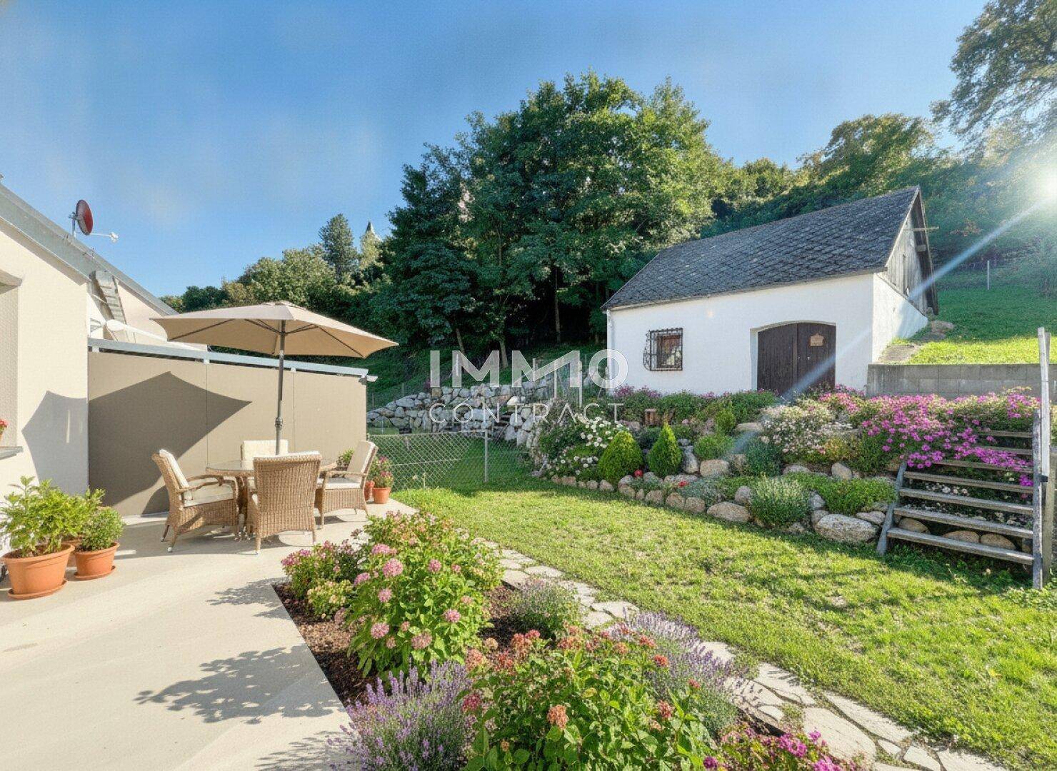 Sonniger Terrassenbereich mit Gartenmöbeln, umgeben von blühenden Beeten und einem charmanten Gartenhaus.