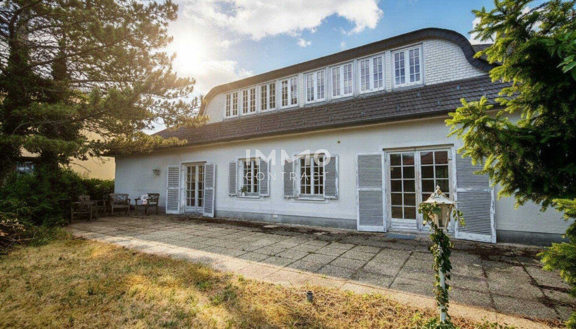 Historische Villa mit Mansarddach, großer Steinterrasse und Gartenbereich mit alten Bäumen.