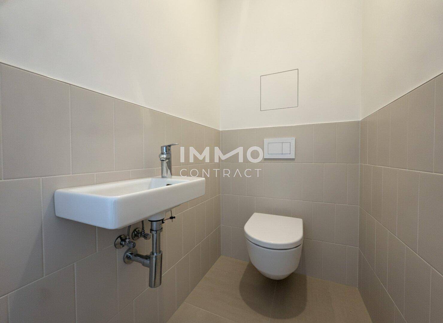 Separates WC mit wandhängender Toilette und kleinem Waschbecken, in hellen Fliesen gehalten.