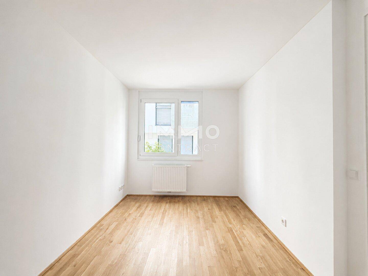 Helles, leeres Schlafzimmer mit Holzfußboden und einem Fenster, das viel Tageslicht hereinlässt.