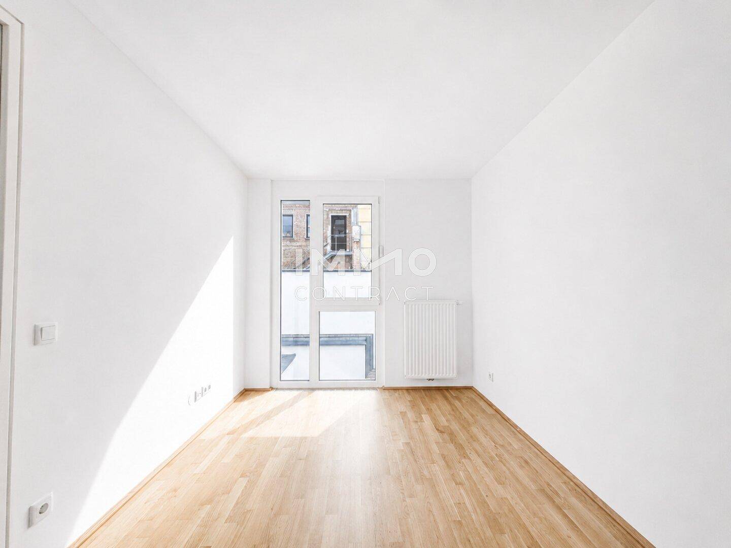 Geräumiges, helles Zimmer mit Holzfußboden und großer Fenstertür, ideal als Schlaf- oder Arbeitszimmer.