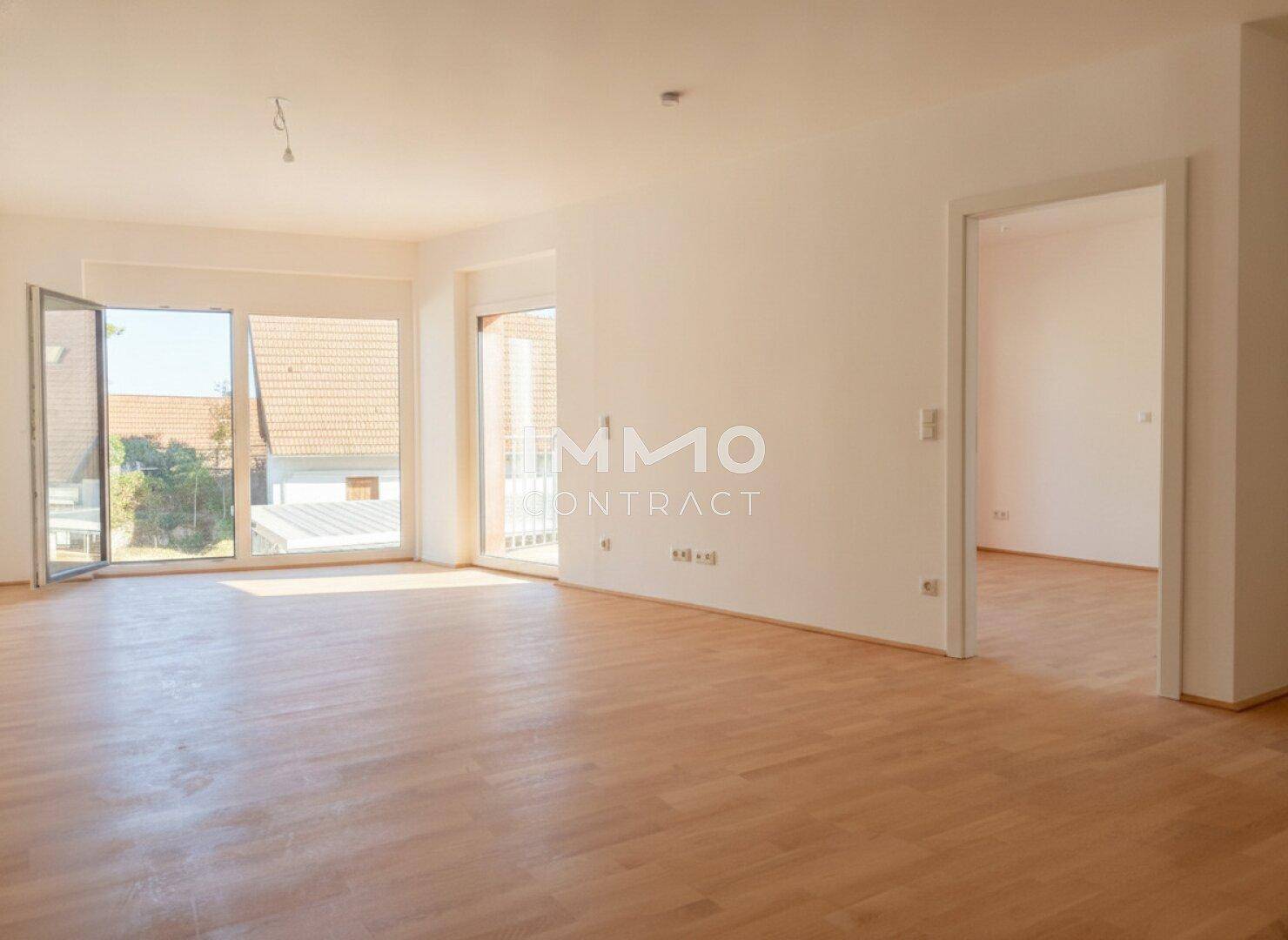 Heller, leerer Wohnraum mit Holzboden und großen Fenstern, die viel Tageslicht hereinlassen und einen Blick ins Grüne bieten.