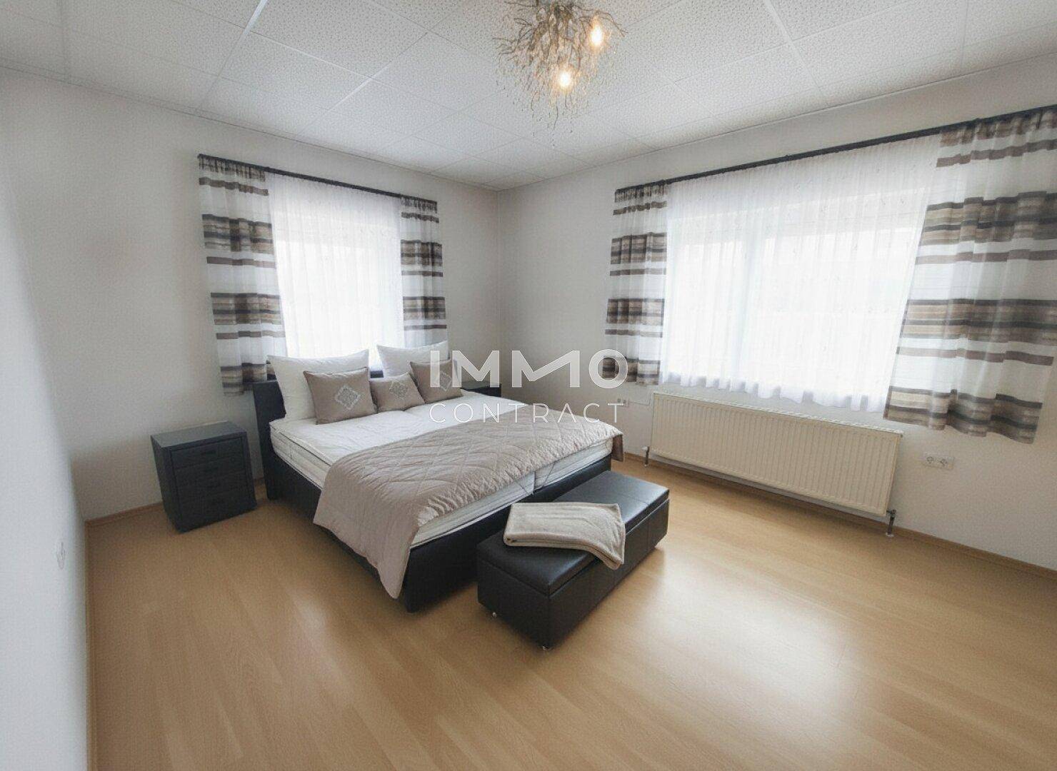 Helles Schlafzimmer mit Doppelbett, Nachttischen, Holzfußboden und großen Fenstern mit Vorhängen.