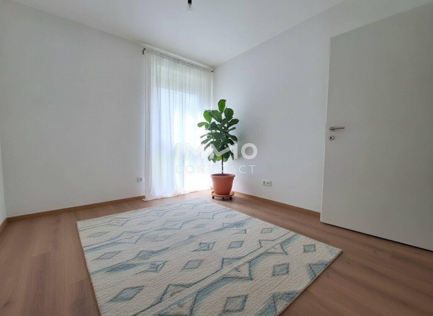 Helles Schlafzimmer mit Laminatboden, großem Fenster, einer Pflanze und einem gemusterten Teppich.