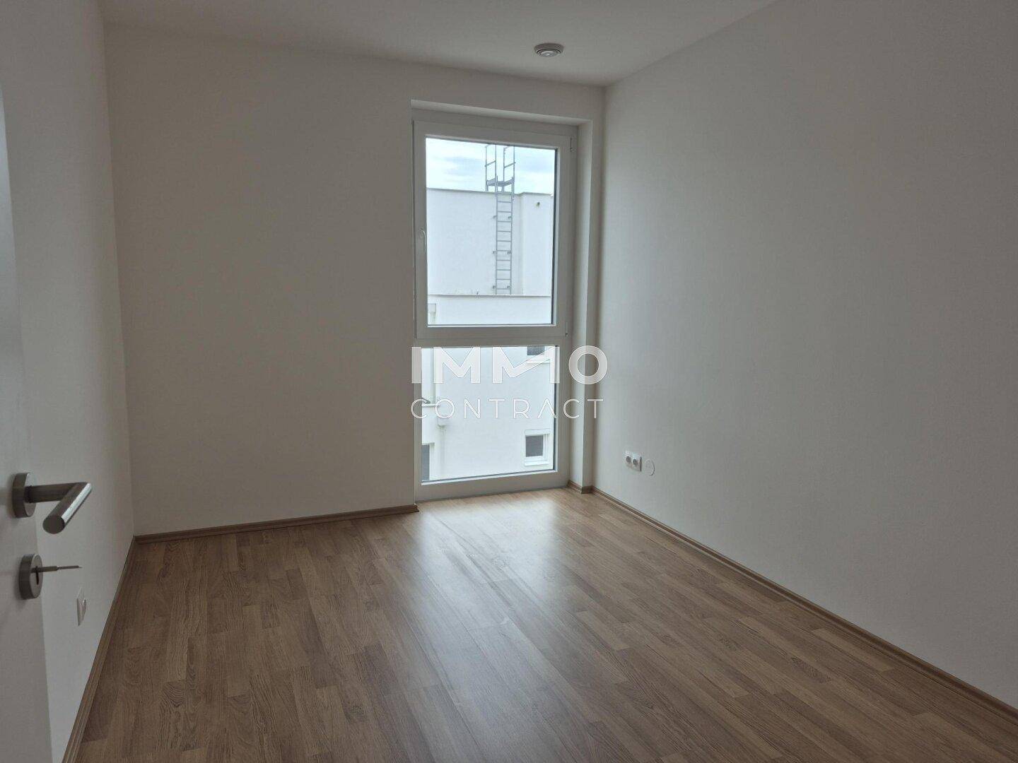 Helles, leeres Zimmer mit Laminatboden und einem großen Fenster, das viel Tageslicht hereinlässt.