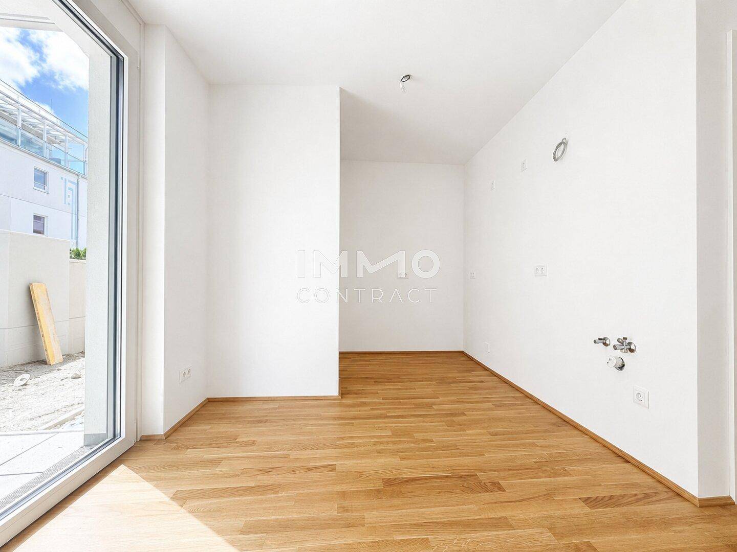 Leerer Raum mit Parkettboden, Küchenanschlüssen und großem Fenster mit Terrassenzugang.