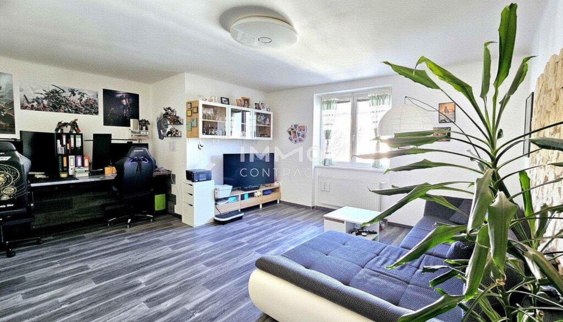 Helles Wohnzimmer mit dunklem Laminatboden, Sofa und integriertem Arbeitsbereich am Fenster.
