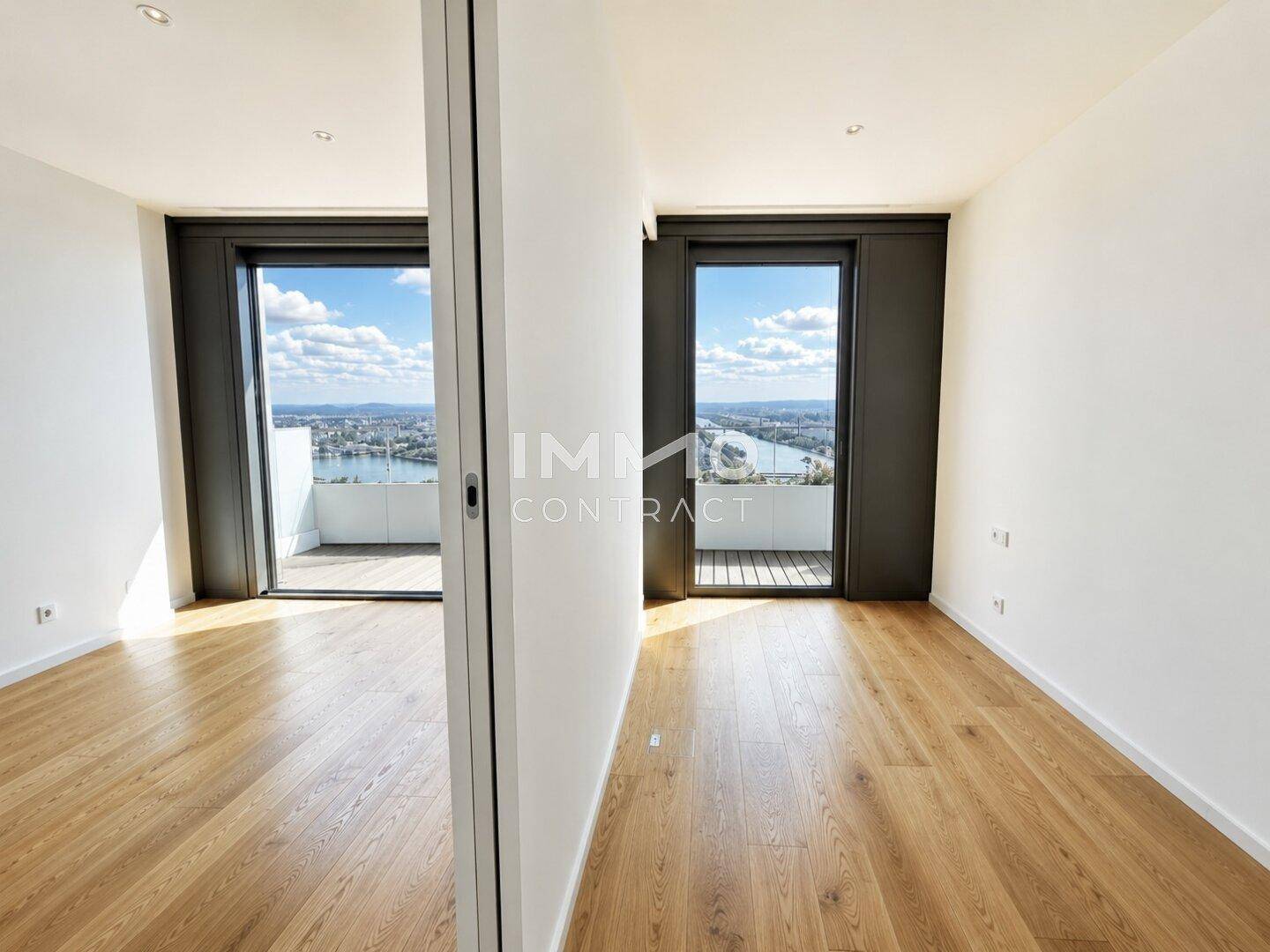 Heller Raum mit Holzboden und großen Fenstern, die Zugang zu einem Balkon bieten und eine weite Aussicht ermöglichen.