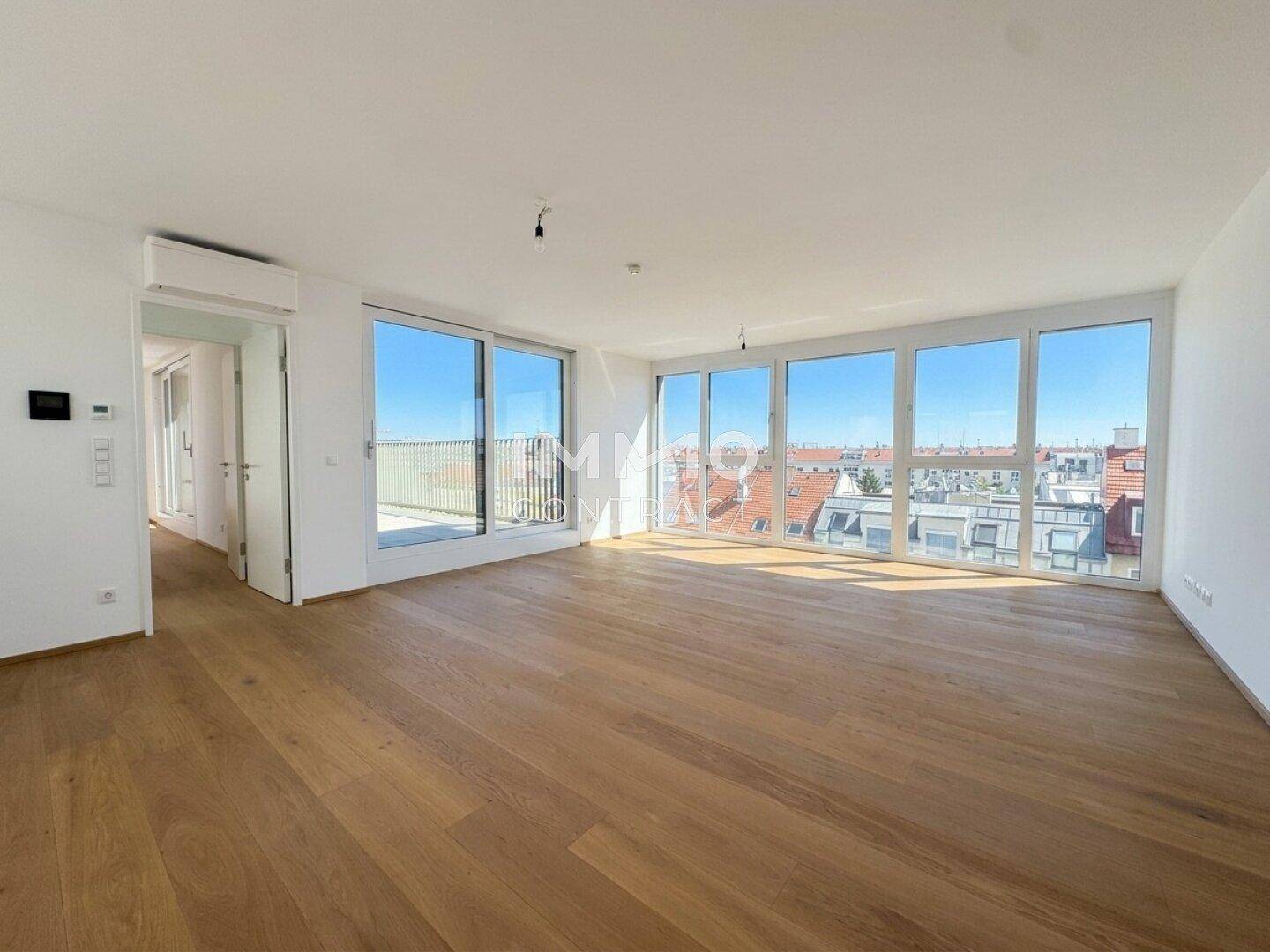Helles, leeres Wohnzimmer mit großen Fenstern, Holzboden und Blick auf die Stadt.