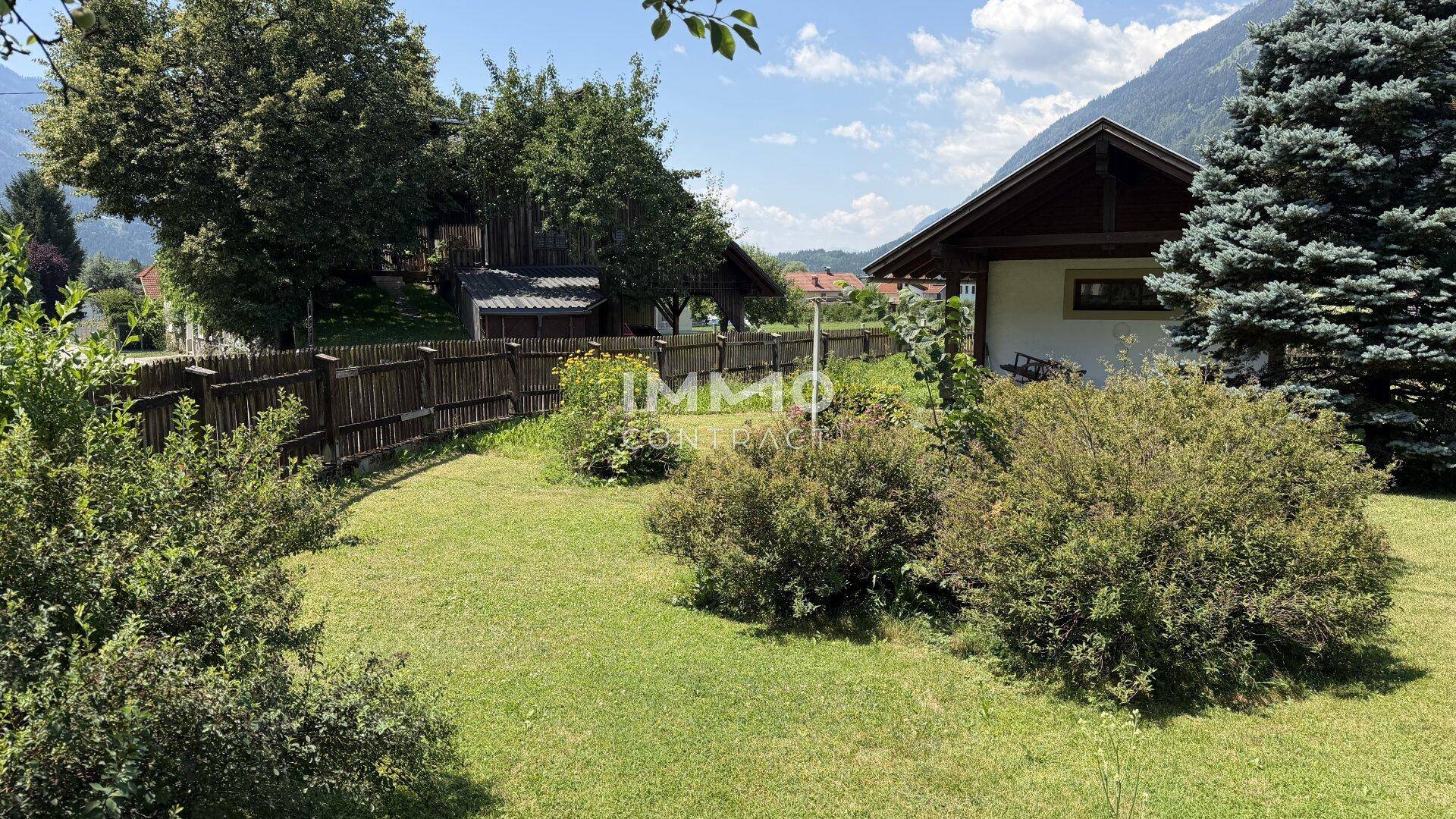 Weitläufiger Gartenbereich mit Rasen, Büschen und Bäumen, umgeben von einem Holzzaun und Bergblick.