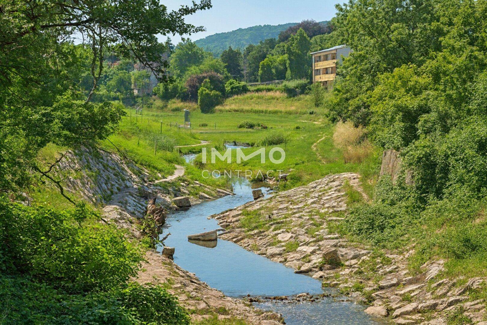 Idyllischer Bachlauf umgeben von üppiger Vegetation und einem Gebäude in der Ferne.