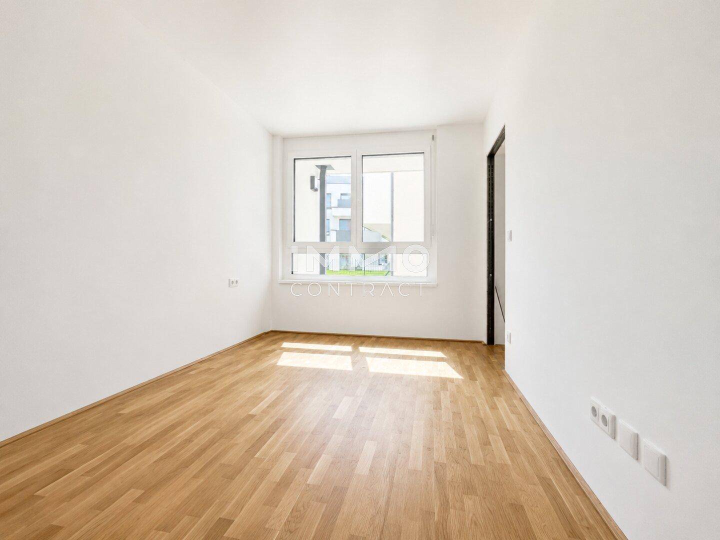 Leeres Zimmer mit hellem Parkettboden und einem großen Fenster, das den Raum mit Licht füllt.
