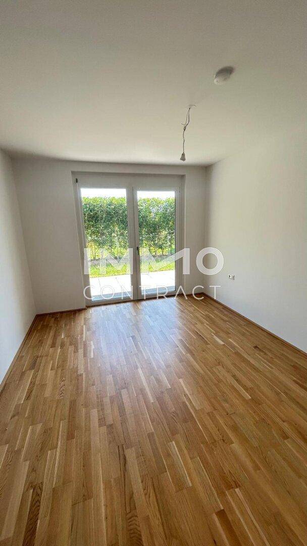 Leeres Zimmer mit Holzboden und Zugang zu einem grünen Außenbereich.
