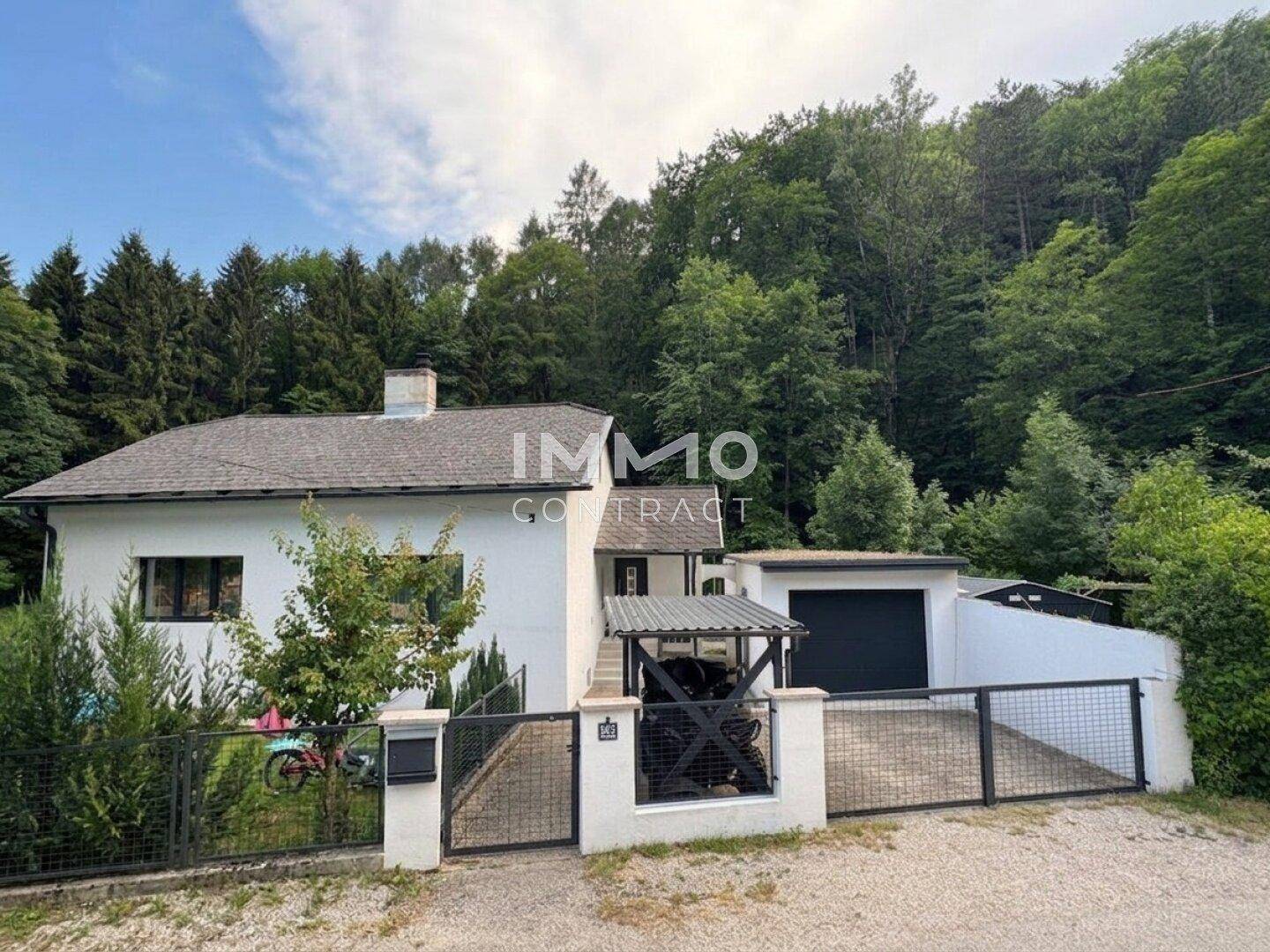 Vorderansicht des Hauses mit Garage, Einfahrt und umzäuntem Vorgarten, umgeben von Bäumen.