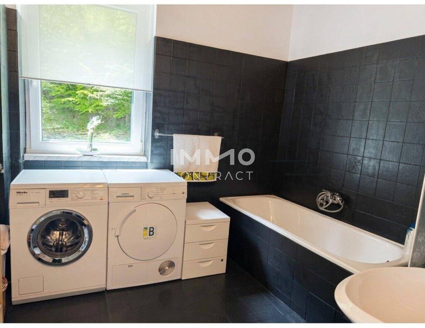 Badezimmer mit schwarzen Fliesen, Badewanne, Waschmaschine und Trockner, sowie einem Fenster.