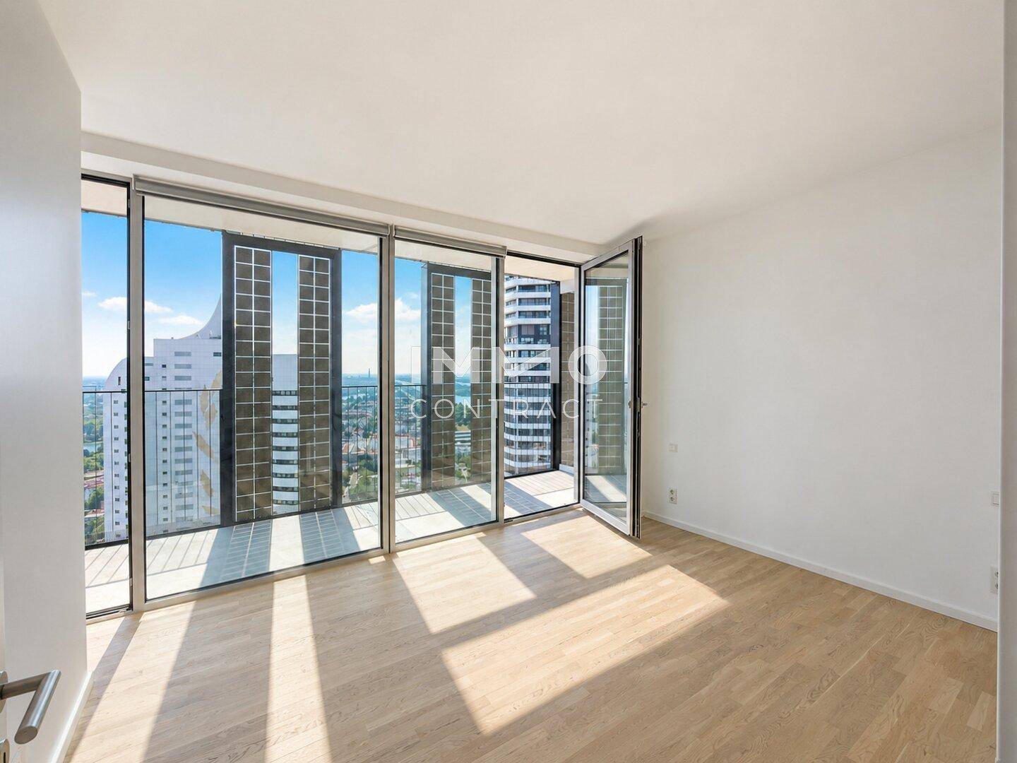 Schlafzimmer mit hellem Parkettboden, großen Fenstern und urbanem Ausblick.