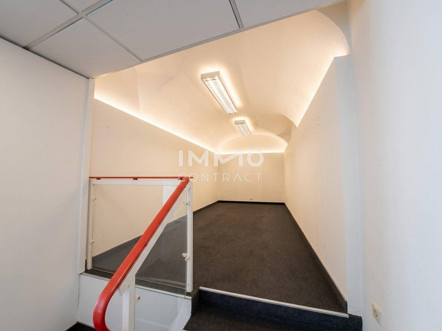 Treppenabgang mit Teppichboden, rotem Handlauf und gewölbter Decke mit Beleuchtung.