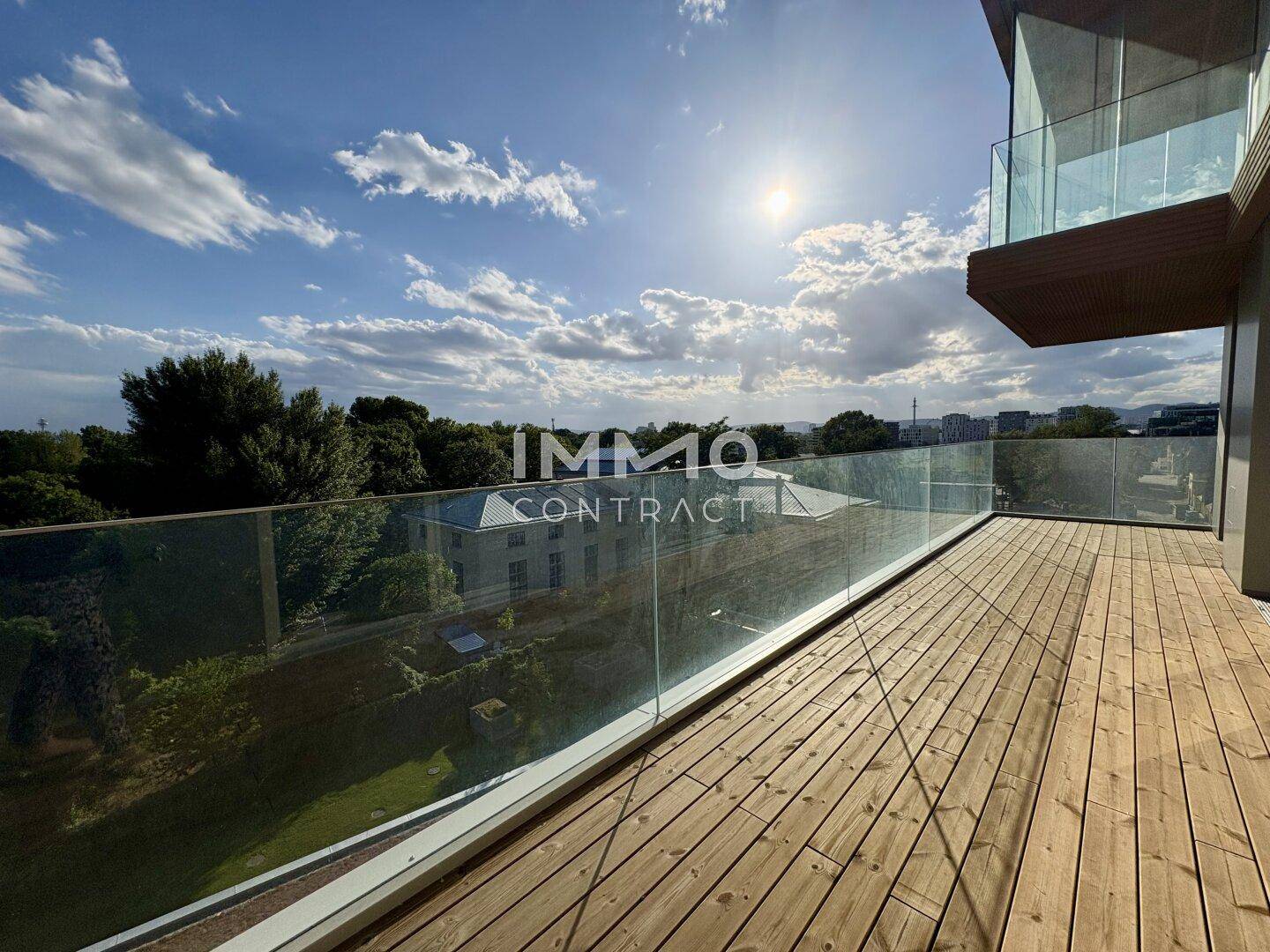 Geräumige Holzterrasse mit Glasgeländer und sonnigem Ausblick ins Grüne.