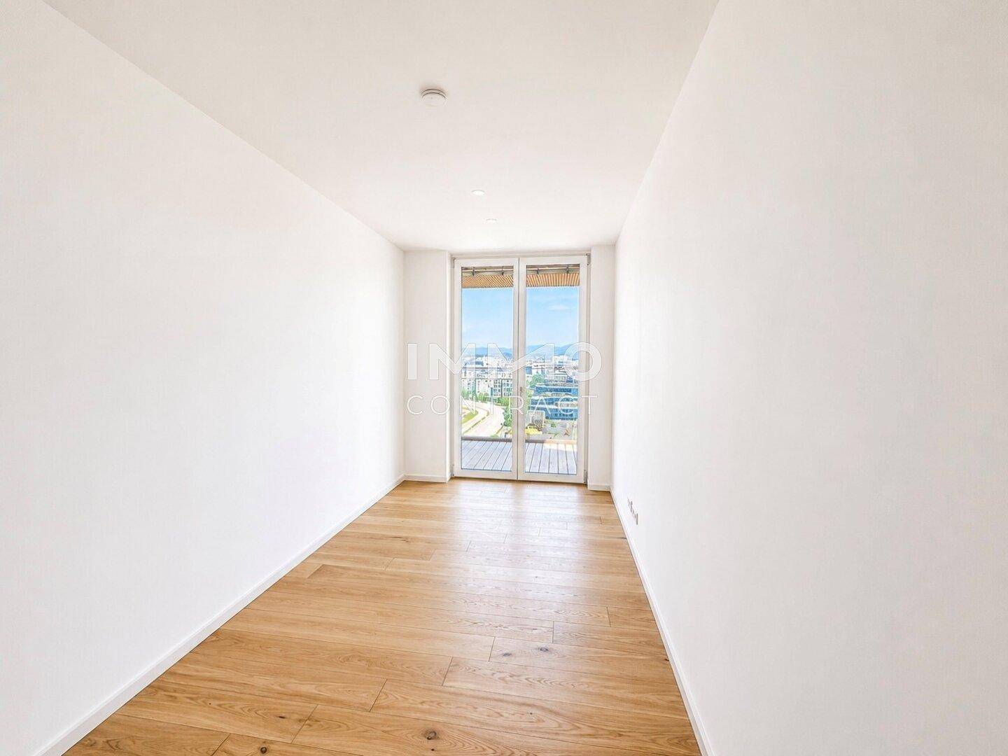 Helles Zimmer mit Holzboden und großen Fenstern, die Zugang zu einem Außenbereich bieten und viel Licht spenden.