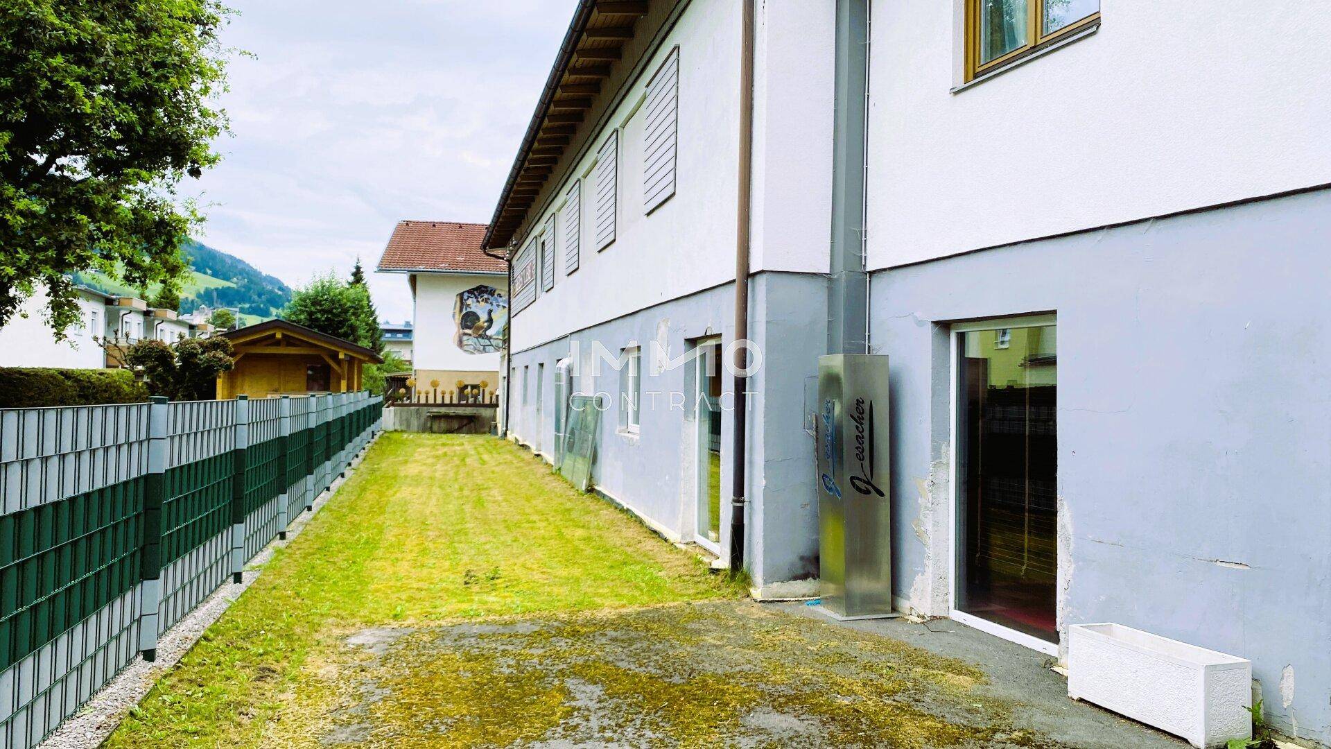 Seitenansicht des Gebäudes mit Rasenfläche, umzäunt von einem grünen Zaun und Bäumen im Hintergrund.