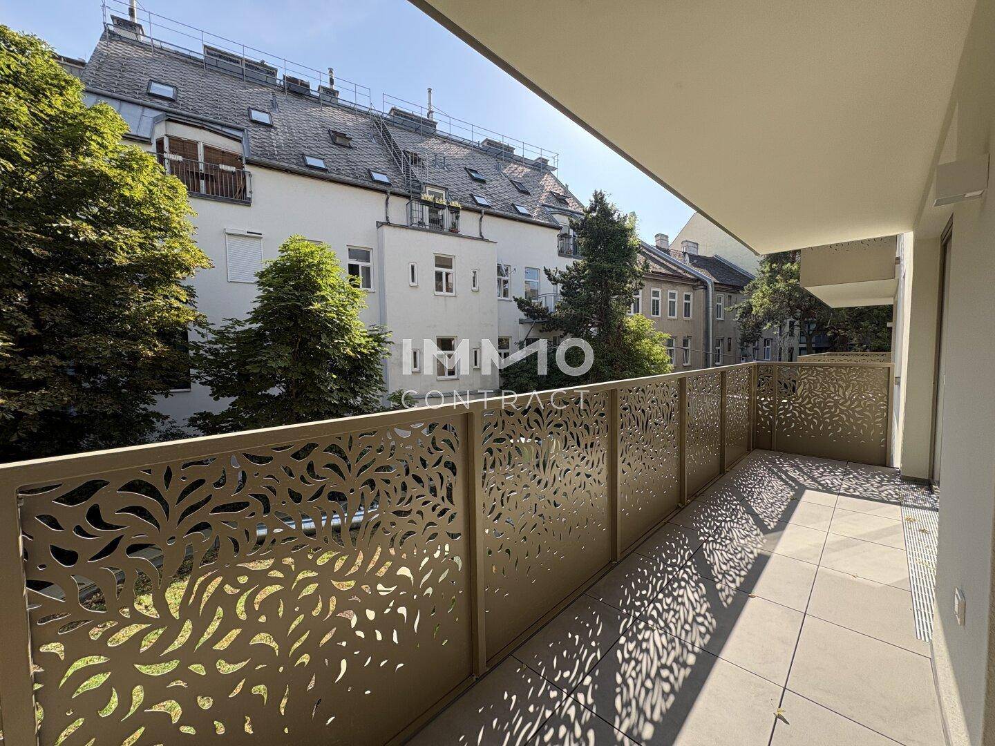 Langer Balkon mit dekorativem Geländer und Fliesenboden, bietet Ausblick auf Bäume und Häuser.