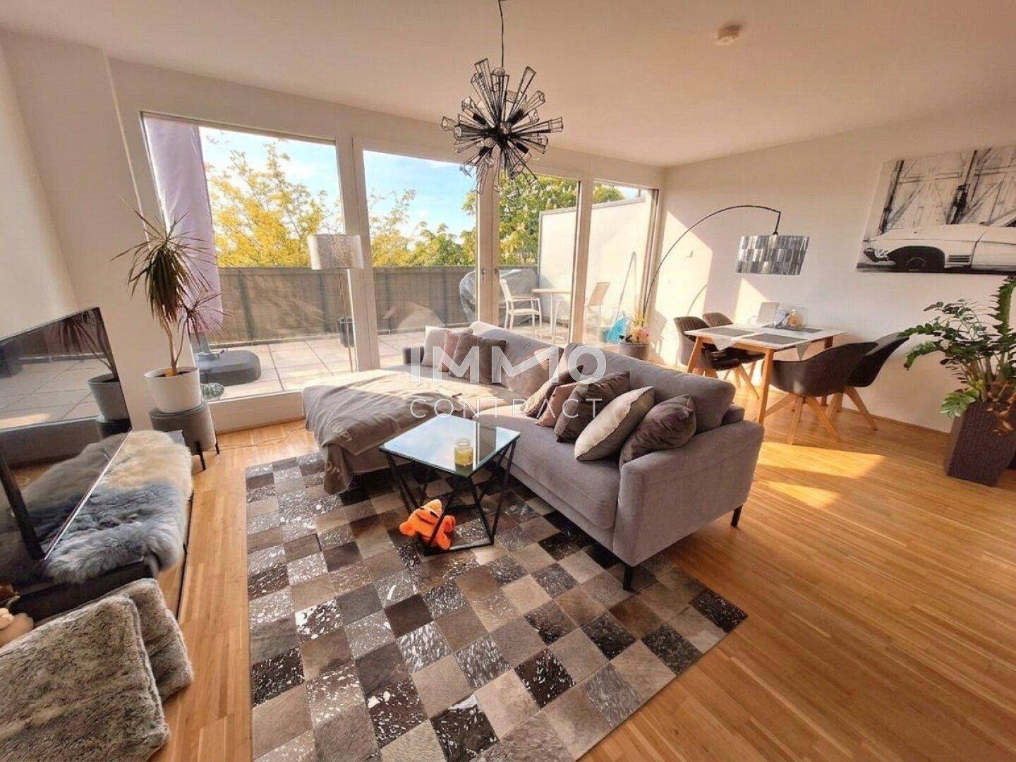 Gemütlicher Wohnbereich mit Sofa, Teppich und Essbereich, hell durch große Fenster.