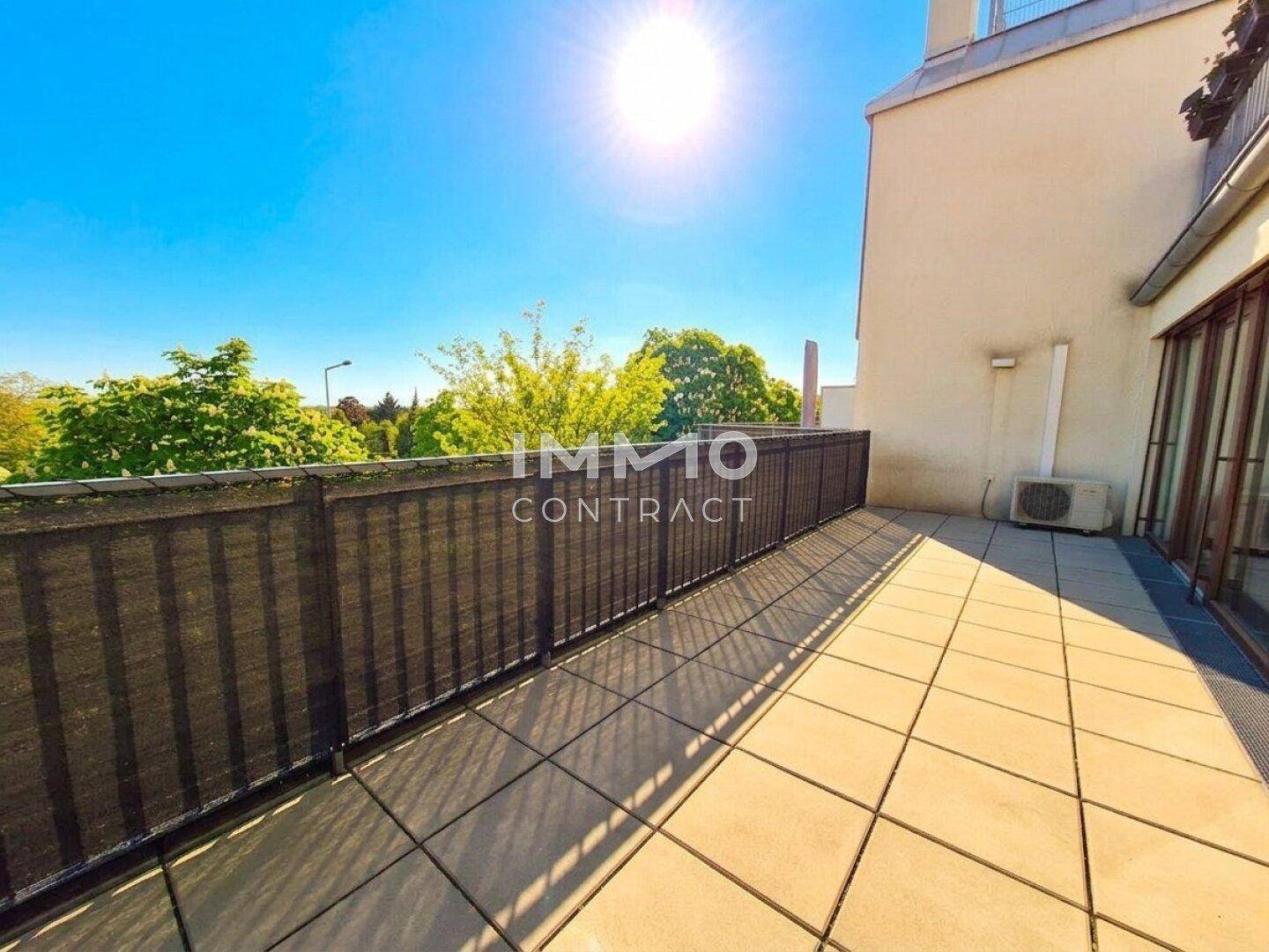 Lange, sonnige Terrasse mit Fliesenboden und Blick auf die grüne Umgebung.