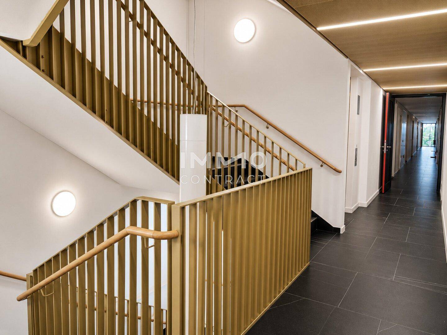 Modernes Treppenhaus mit Holzgeländer und dunklen Fliesen in einem Mehrfamilienhaus.
