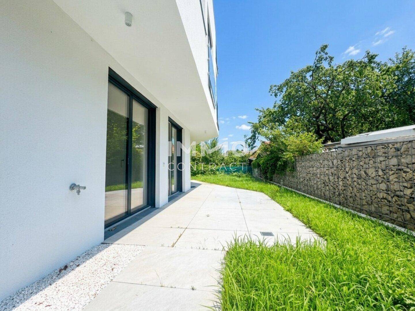 Langer, gefliester Terrassenbereich entlang des Gebäudes mit Gabionenwand und Rasenfläche.