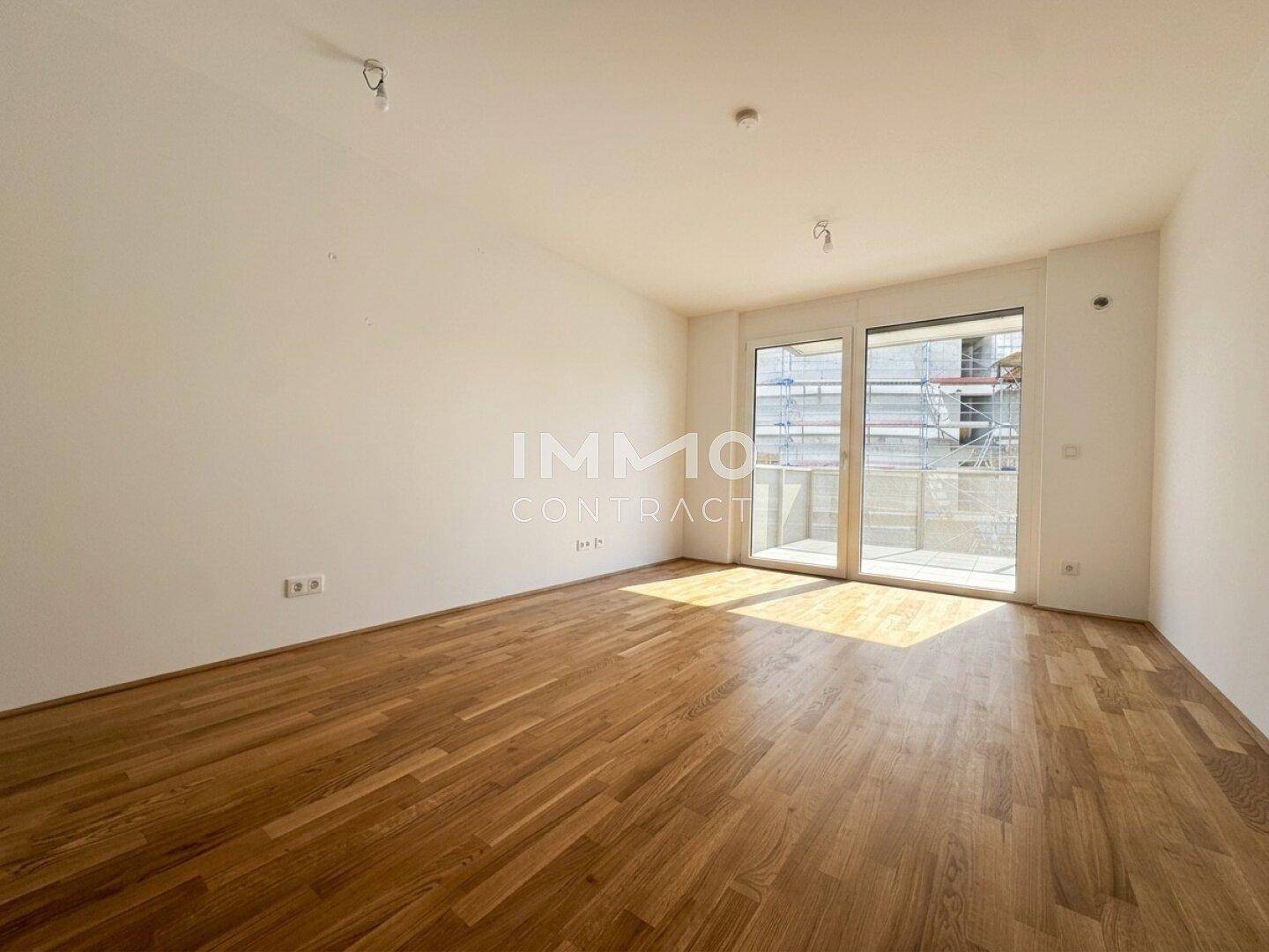 Heller, leerer Raum mit Holzparkettboden und großem Fenster, das viel Tageslicht hereinlässt.