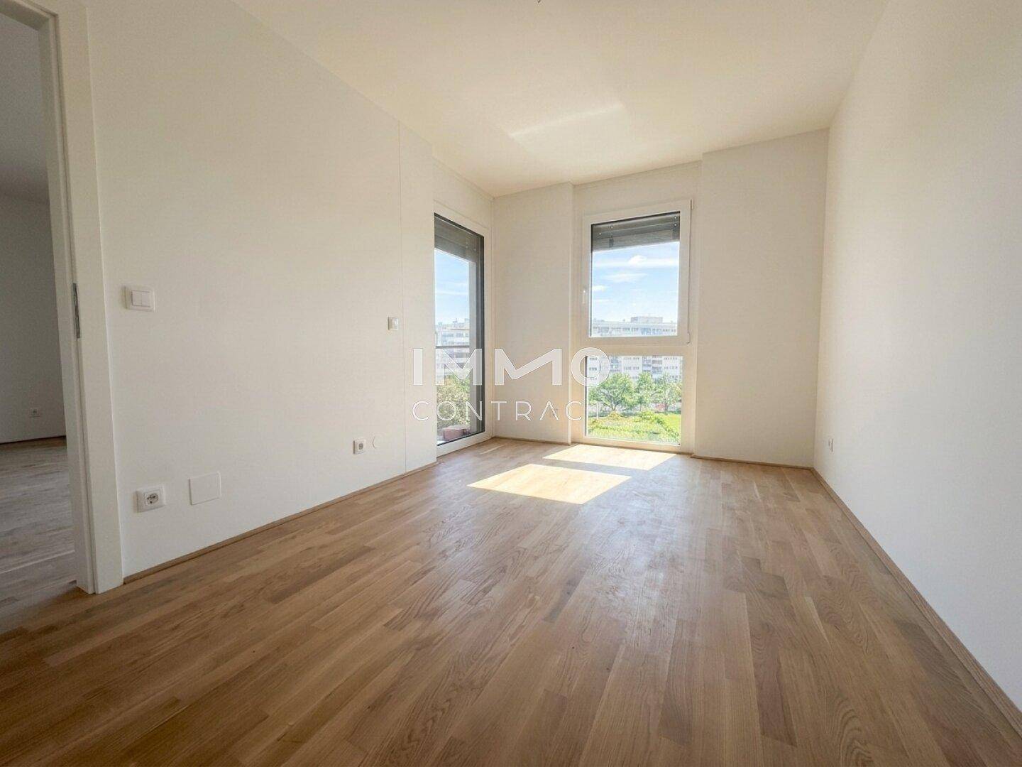 Helles Schlafzimmer mit Parkettboden und zwei Fenstern, die einen Blick ins Grüne bieten.