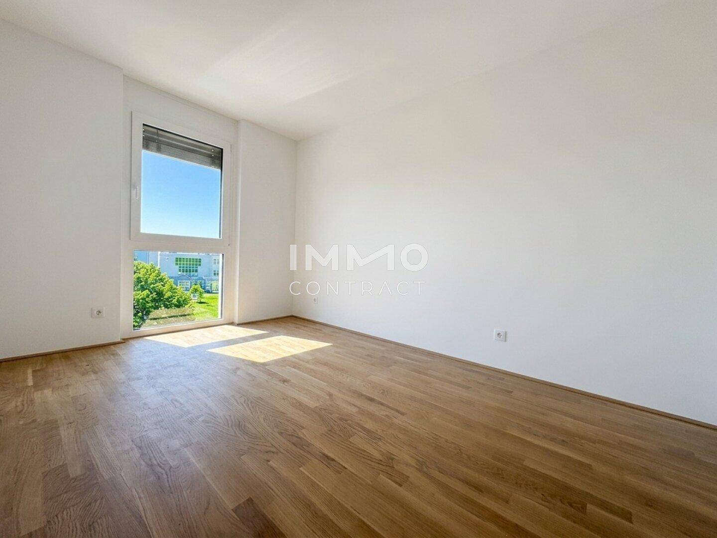Leeres Zimmer mit hellem Parkettboden und einem großen Fenster, das viel Licht spendet.