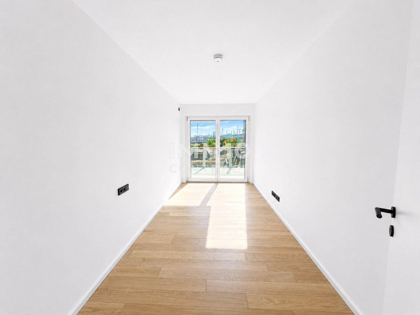 Helles, leeres Zimmer mit Holzboden und Balkonzugang durch eine große Glastür.