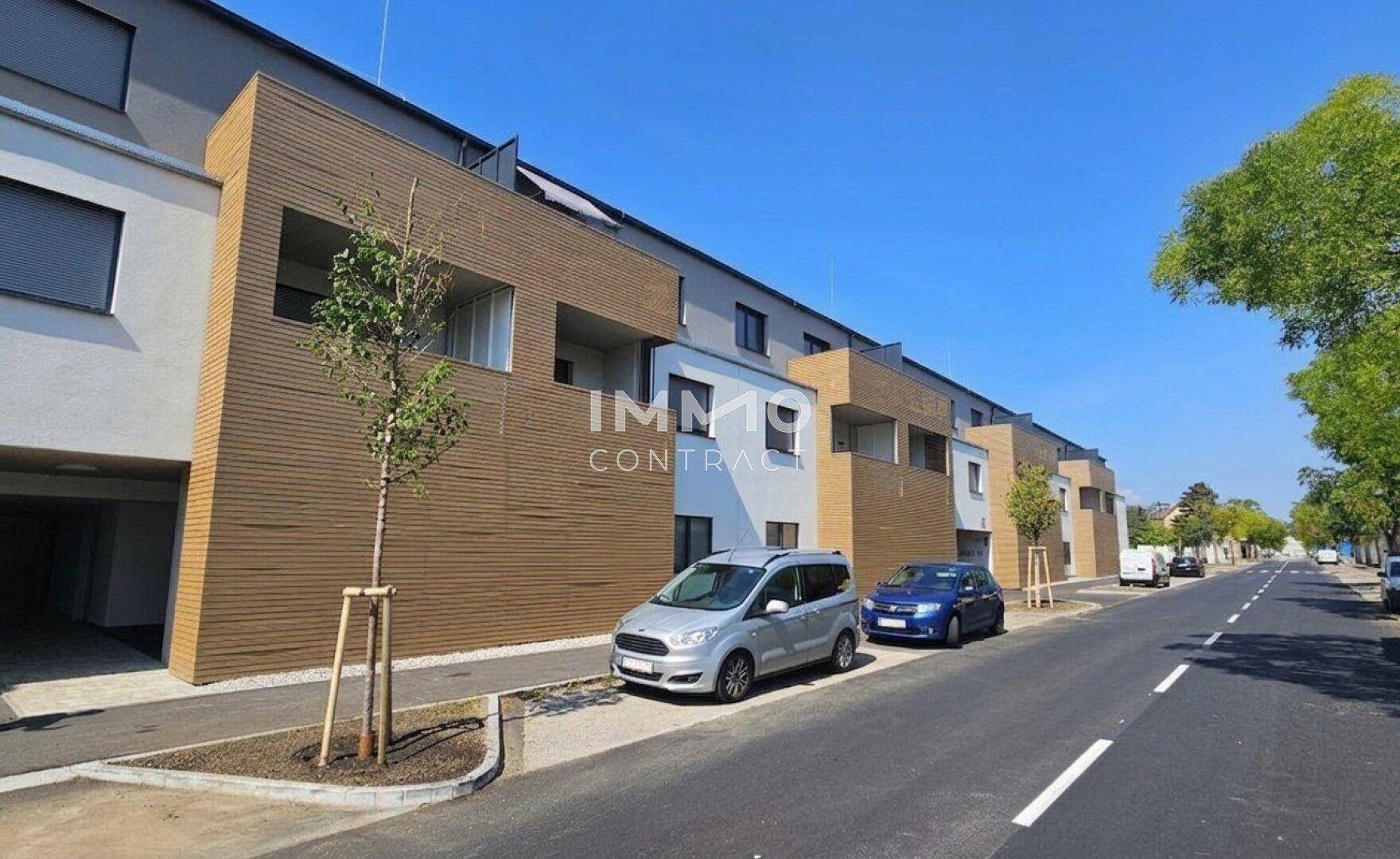 Ansicht der modernen Wohnanlage von der Straße mit geparkten Fahrzeugen und Bäumen.