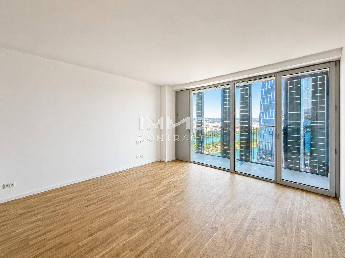 Geräumiges, leeres Zimmer mit Holzboden und großen Fenstern, die einen weiten Stadtblick ermöglichen.