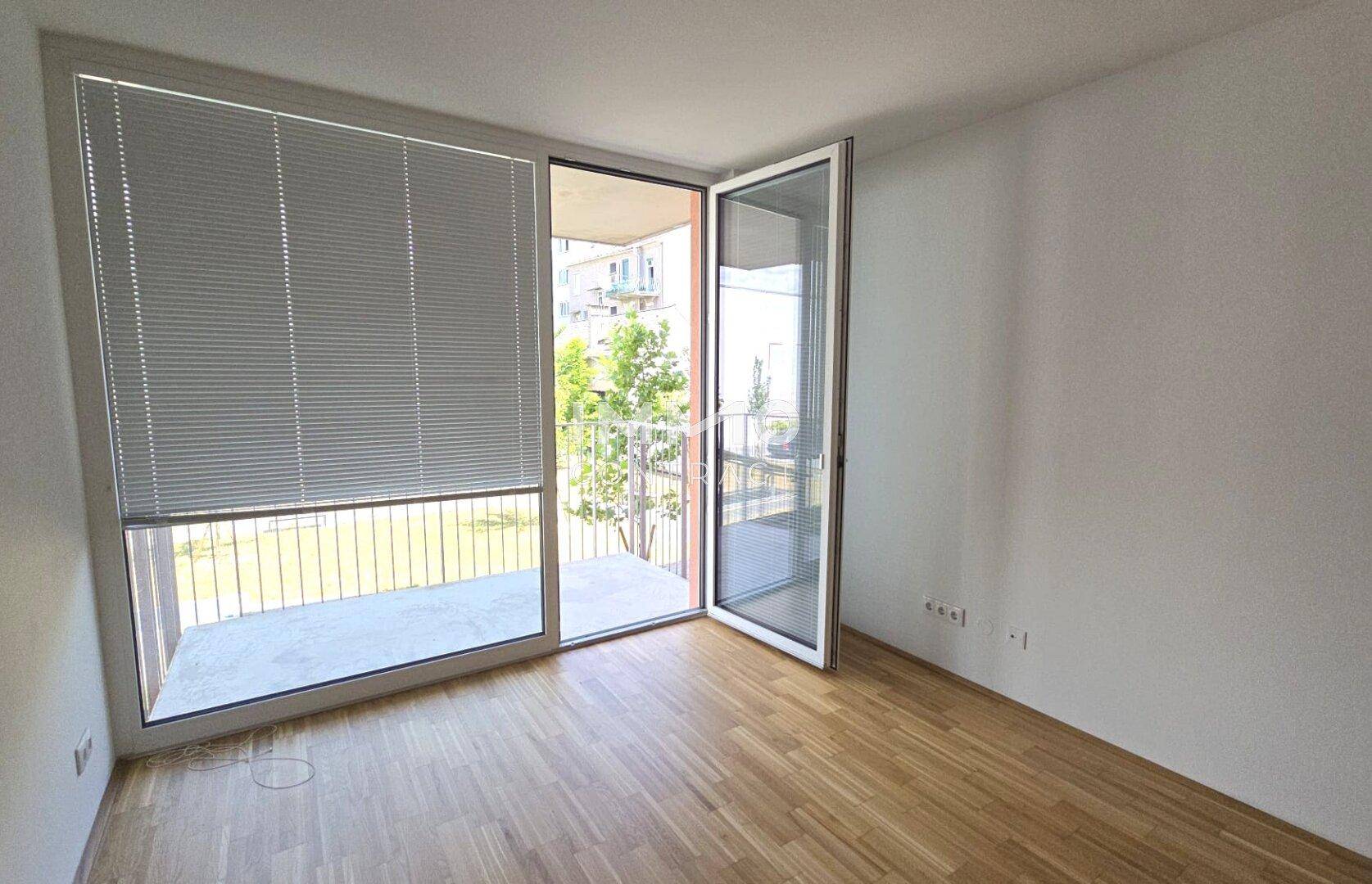 Helles Zimmer mit Parkettboden und direktem Zugang zu einem Balkon, ausgestattet mit Jalousien.