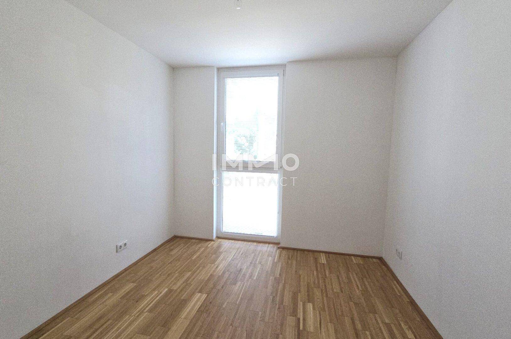Helles Zimmer mit Parkettboden und einem hohen Fenster, das viel Tageslicht hereinlässt.